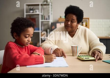 Black senior woman assisting Black child with homework at table, child writing in notebook while woman pointing at page, calculator and striped mug on desk, home setting Stock Photo
