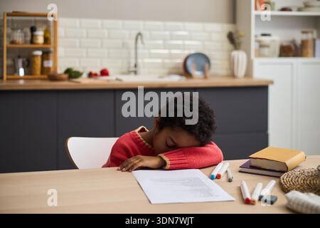 Black child resting head on table while sitting in kitchen, appearing tired or bored, surrounded by scattered markers, paper, and books, suggesting homework or studying activity Stock Photo