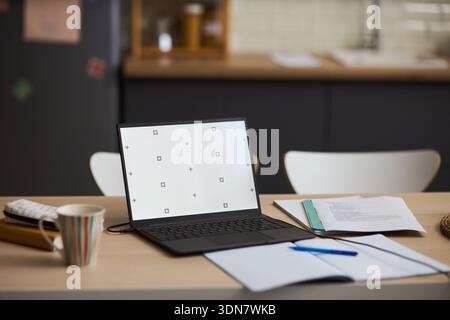Open laptop with blank screen standing on kitchen table next to open notebook, pen, calculator and documents, suggesting remote work or study environment in modern home interior Stock Photo