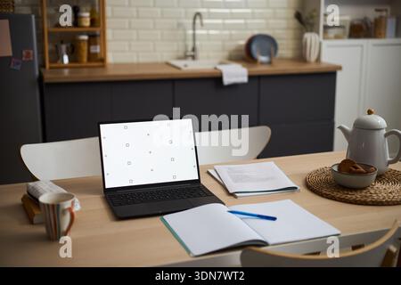 Modern kitchen table featuring open laptop with blank screen, scattered notebooks and pen, teapot and cup on woven mat, suggesting remote work or study environment Stock Photo