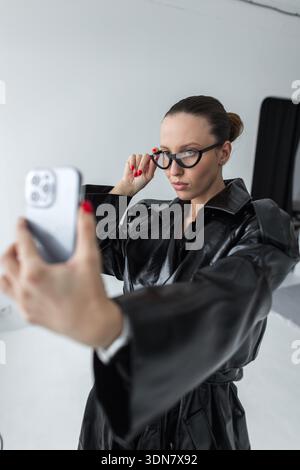 Stylish woman in elegant black outfit and designer eyewear using a smartphone for a selfie indoors, symbolizing self-expression, digital communication Stock Photo