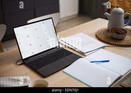 Open laptop with blank screen standing on wooden table next to open notebook, pen, documents and cup, showing workspace setup for studying, working or remote learning at home Stock Photo