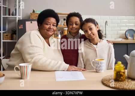 Middle aged Black woman sitting at kitchen table with two Black children, smiling and embracing them while looking at camera, paper and mugs visible on table in foreground Stock Photo