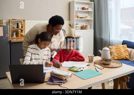 Middle aged Black woman assisting two Black children with homework at kitchen table, children writing in notebooks, laptop and school supplies on table, family learning together Stock Photo