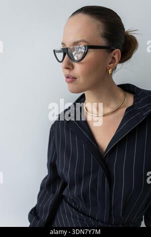 Close-up of a stylish businesswoman in a pinstripe shirt and trendy black glasses. Elegant corporate look with gold jewelry and a sleek bun against a Stock Photo