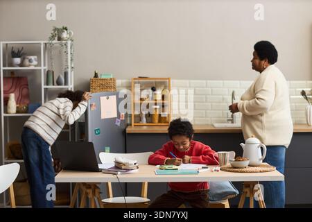 Black boy sitting at table drawing with colored pencils, Black woman standing at kitchen counter preparing food, Black teenager reaching for item on shelf in modern kitchen Stock Photo