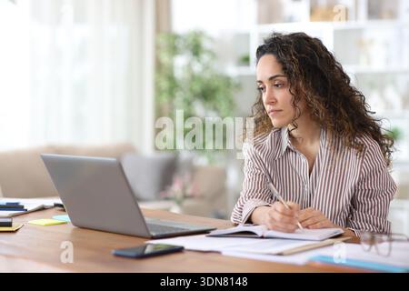 Adult freelance worker taking note in agenda checking laptop working at home Stock Photo
