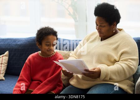 Black senior woman sitting on sofa showing documents to Black child, both engaging in conversation, child looking at papers with interest, home environment visible in background Stock Photo