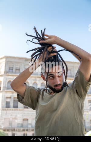 Young man with dreadlocks, holding his hair against a city building background Stock Photo