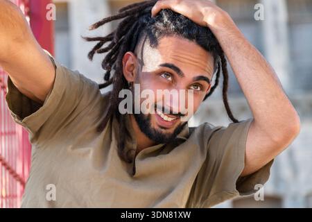 Smiling young man touching his dreadlocks near a red fence on a sunny day Stock Photo