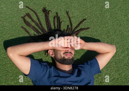 Young man with dreadlocks lying on the green grass, dazzled by the sun Stock Photo