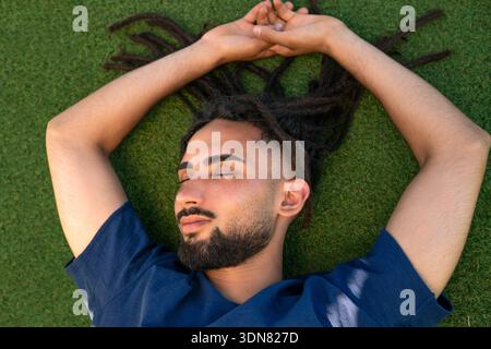 Young man with dreadlocks lying on green grass, feeling peaceful Stock Photo