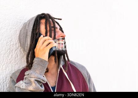 Young man with dreadlocks smiling on the phone against a white wall Stock Photo