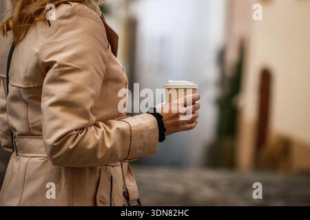 Woman in trench coat holding paper coffee cup walking on city street. Take-away hot drink Stock Photo