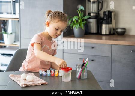 Child dipping brush into jar of paint for egg decoration. Ritualistic preparation for festive activity. Stock Photo