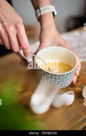 Hand mixing beaten eggs in ceramic bowl on kitchen counter with eggshells and wooden cutting board Stock Photo
