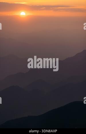 Sun setting behind the misty silhouette of layered mountain ridges under a golden sky. Mountains of Ta Xua, Bac Yen District, Son La, Vietnam Stock Photo