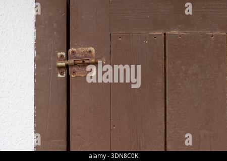 Close-up of an aged Rusty Metal Slide Bolt Lock securing a weathered wooden door beside a white stucco wall, highlighting rustic textures, security, p Stock Photo