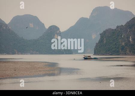 A lone boat on a vast, serene lake surrounded by towering green hills under an overcast sky. Cat Ba, Vietnam Stock Photo