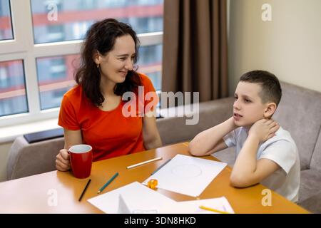 Mother Helping Son with Homework at Home. Mother and son sit at a table with papers and pencils, having a conversation during a study session in a Stock Photo