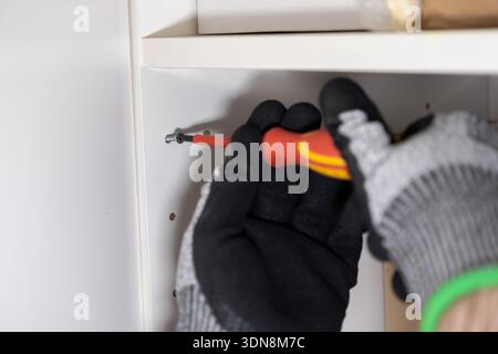 Close-up of a person's gloved hand using a red and yellow screwdriver to meticulously install a screw into a white cabinet panel, emphasizing home imp Stock Photo