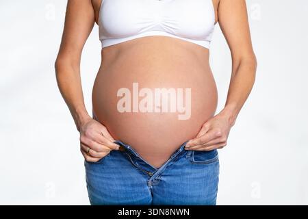 Front view of a pregnant woman with a big belly trying to put on a trouser on white background Stock Photo