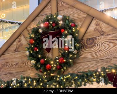 A traditional Christmas wreath, decorated with red and silver ornaments and bright lights, hangs on a rustic wooden wall, symbolizing festive holiday Stock Photo