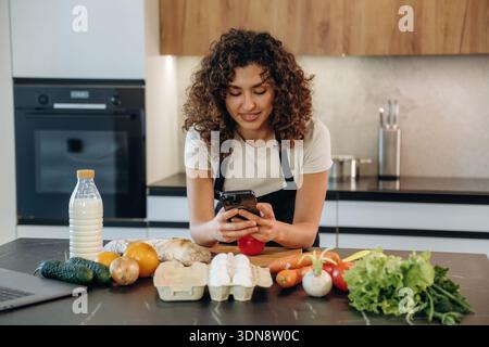 The smartphone is in hands. Attractive woman preparing fresh vegetables on the kitchen counter during cooking. Stock Photo