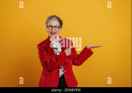 Elderly woman with short gray hair and glasses is smiling broadly while pointing at an empty area against a bright yellow backdrop, radiating joy and Stock Photo