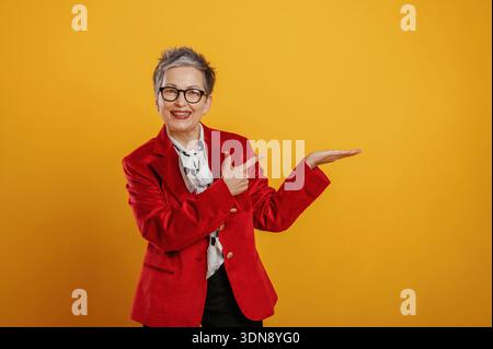 Older woman with short gray hair, wearing a red blazer, is smiling and pointing with her hand against a bright yellow background, creating a lively an Stock Photo