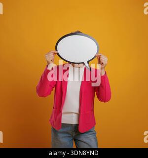 Female figure dressed in a bright pink cardigan is holding a speech bubble in front of her face against a vivid yellow backdrop, symbolizing communica Stock Photo