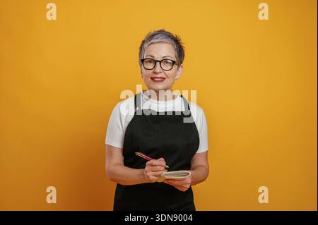 Smiling mature woman with short gray hair and glasses is holding a notebook and pen while wearing a black apron against a bright yellow background, ra Stock Photo