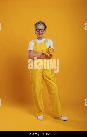Elderly woman in bright yellow overalls and glasses stands with arms crossed, smiling warmly against a vivid yellow backdrop, radiating positivity and Stock Photo