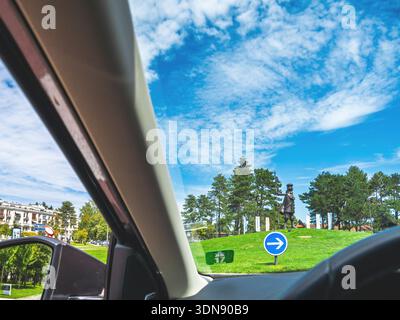 Le Touquet, France - Aug 22, 2023: POV from car windshield showing traffic circle with blue arrow sign, sculpture monument on grass mound, pine trees Stock Photo