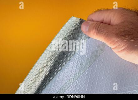 Person peeling and handling transparent plastic bubble wrap sheet used for protective packaging with air-filled bubbles visible against bright orange backdrop. Stock Photo