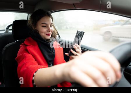 caucasian woman sits in a car, distracted and looking at a smartphone message in her hands, illustrating the danger of texting while driving. Stock Photo