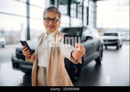 Confident senior woman dressed in a stylish outfit is smiling while holding a credit card in a modern car dealership, surrounded by various vehicles, Stock Photo