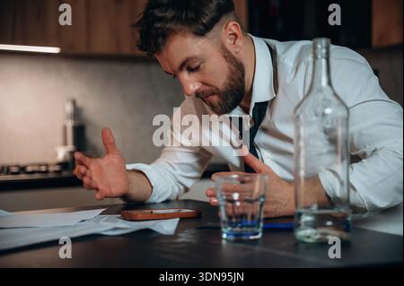 Caucasian man in a white shirt and loosened tie is seated at a desk, surrounded by empty alcohol bottles and papers, showing signs of stress and deep Stock Photo