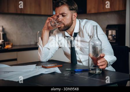 Caucasian man in white shirt and black tie sits at a kitchen table, holding a glass of whiskey, surrounded by scattered papers and an empty bottle, re Stock Photo