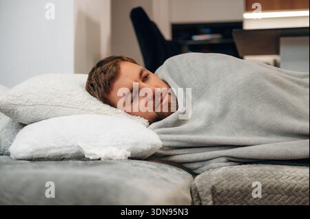 Relaxed young man with light brown hair is lying on a comfortable couch, wrapped in a soft gray blanket, surrounded by fluffy pillows, creating a sere Stock Photo