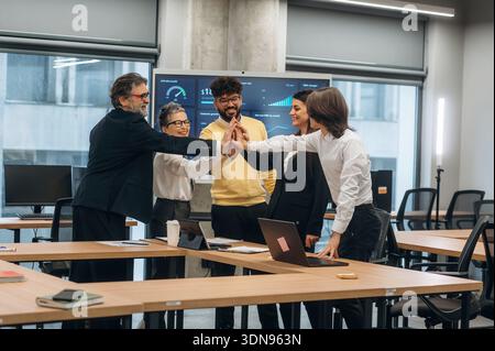 Diverse team of business professionals engaging in a high-five celebration in a contemporary office space, surrounded by technology and collaborative Stock Photo