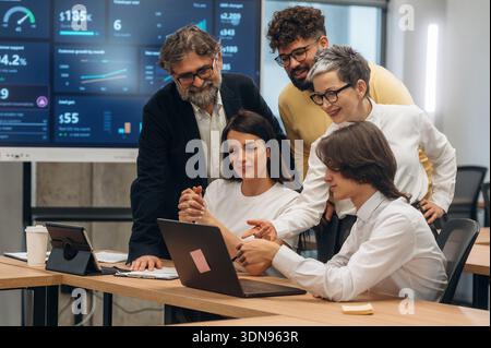Diverse team of business professionals engaged in collaboration within a contemporary office, analyzing data displayed on a laptop, emphasizing teamwo Stock Photo