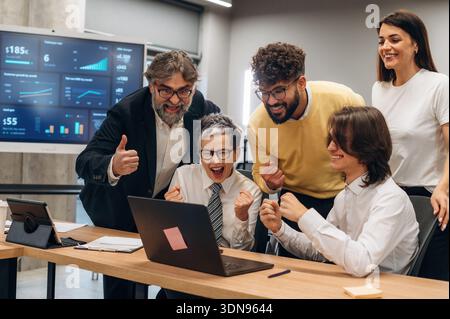 Diverse team of business professionals joyfully celebrating a successful project in a contemporary office, gathered around a laptop displaying perform Stock Photo