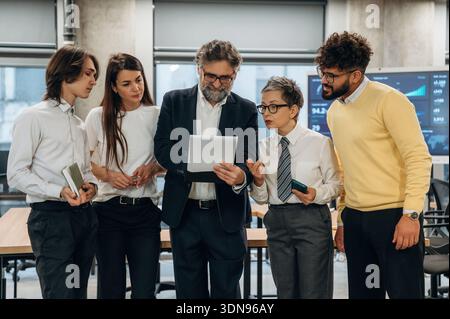 Diverse team of business professionals engaged in a collaborative discussion in a modern office, analyzing data on a digital tablet, highlighting team Stock Photo