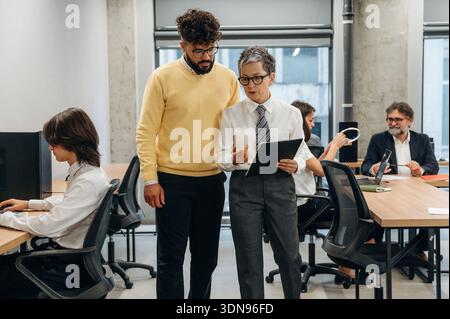Two business professionals engage in a discussion about project details in a contemporary office setting, with colleagues focused on their tasks at de Stock Photo