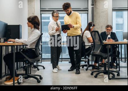 Two business professionals are collaborating in a modern office space, discussing project details while utilizing a digital tablet, surrounded by coll Stock Photo