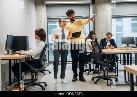 Two business professionals engage in a lively discussion in a contemporary office space, surrounded by colleagues focused on their tasks, highlighting Stock Photo