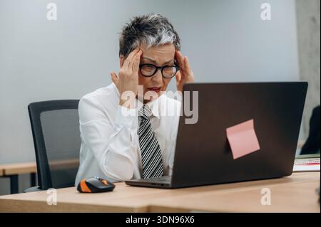 Businesswoman with short gray hair is visibly stressed while working on her laptop in a contemporary office, highlighting the pressures of corporate l Stock Photo