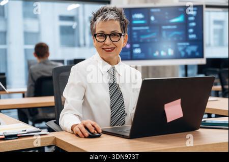 Confident businesswoman with short gray hair is smiling while using a laptop in a modern office, featuring collaborative workspaces, technology, and d Stock Photo