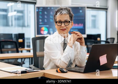 Confident businesswoman with short gray hair is seated at a sleek desk in a modern office, smiling at the camera while surrounded by digital displays Stock Photo
